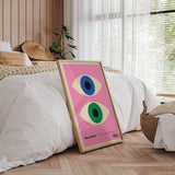 Bauhaus plakat med to øjne i forskellige farver på en lyserød baggrund, placeret i en ramme ved siden af en seng i et moderne soveværelse.