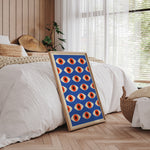 Plakat med geometrisk mønster i blå, rød og orange, placeret på gulvet i et soveværelse med træpaneler og naturligt lys.