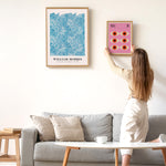 Kvinde hænger en plakat op på en hvid væg ved siden af en sofa. På væggen hænger allerede en William Morris plakat med blomstermotiv.