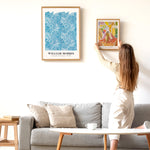 En kvinde hænger en plakat op på en hvid væg over en grå sofa. Den ene plakat er af William Morris, og den anden er "Faun og Nymf".