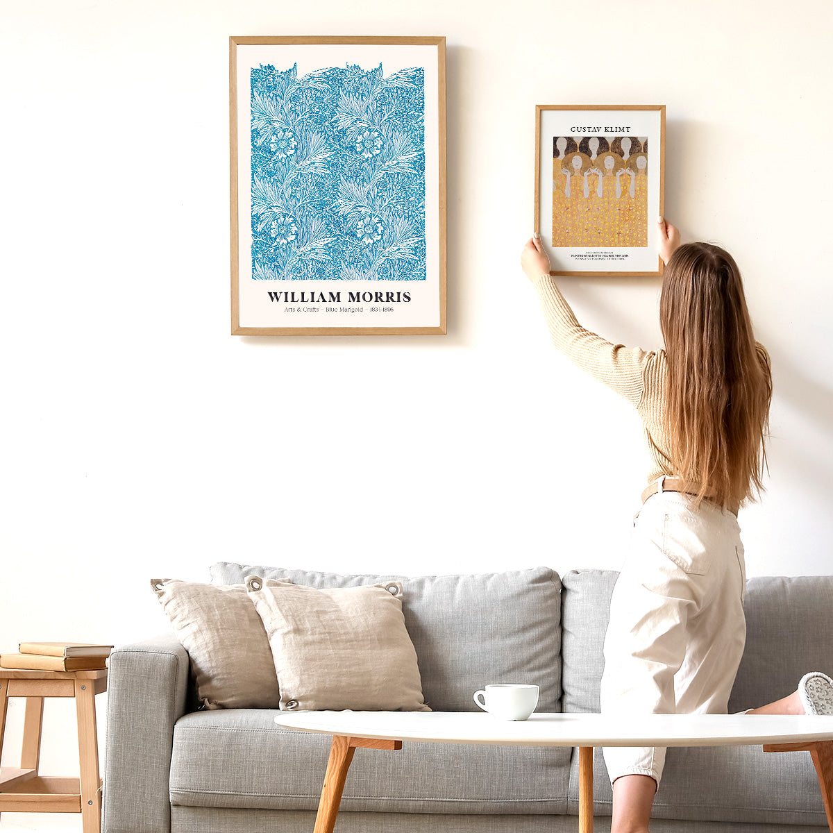Kvinde hænger en Gustav Klimt-plakat op på en væg ved siden af en William Morris-plakat i en stue.