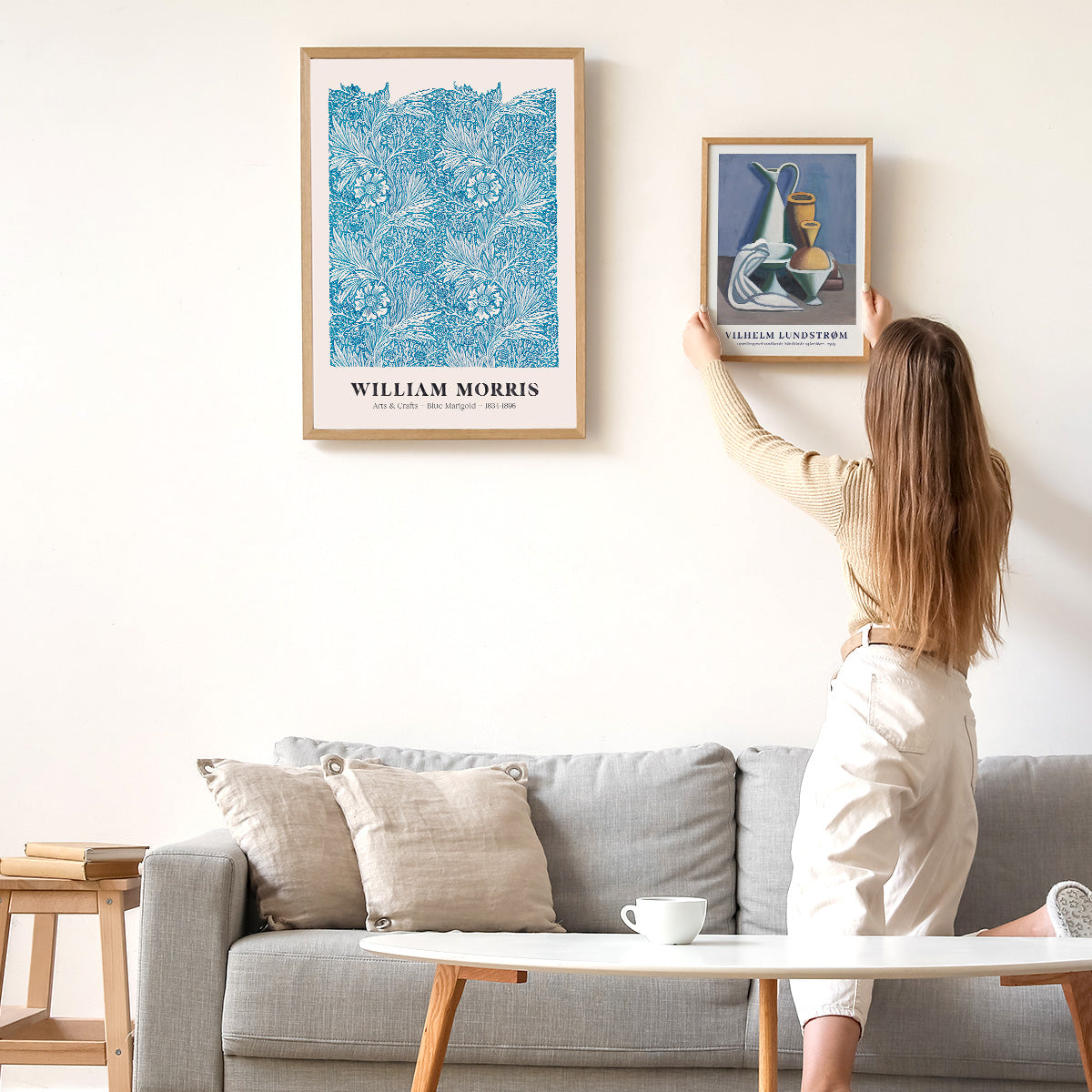 Kvinde hænger en plakat op på en hvid væg over en grå sofa. Plakaten til venstre er af William Morris med et blåt blomstermønster.