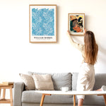 Kvinde hænger en plakat op på en hvid væg over en grå sofa. Den venstre plakat er af William Morris med et blåt blomstermønster.