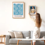 Kvinde hænger en plakat op på en væg ved siden af en William Morris plakat. Plakaterne er indrammet og hænger over en grå sofa i en moderne stue.
