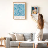 Kvinde hænger en plakat op på en væg ved siden af en William Morris plakat. Plakaterne er indrammet og hænger over en grå sofa i en moderne stue.