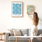 Kvinde hænger en William Morris plakat op på en væg over en sofa. En anden William Morris plakat er allerede hængt op.