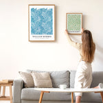 Kvinde hænger William Morris-plakat op på væggen over en grå sofa.