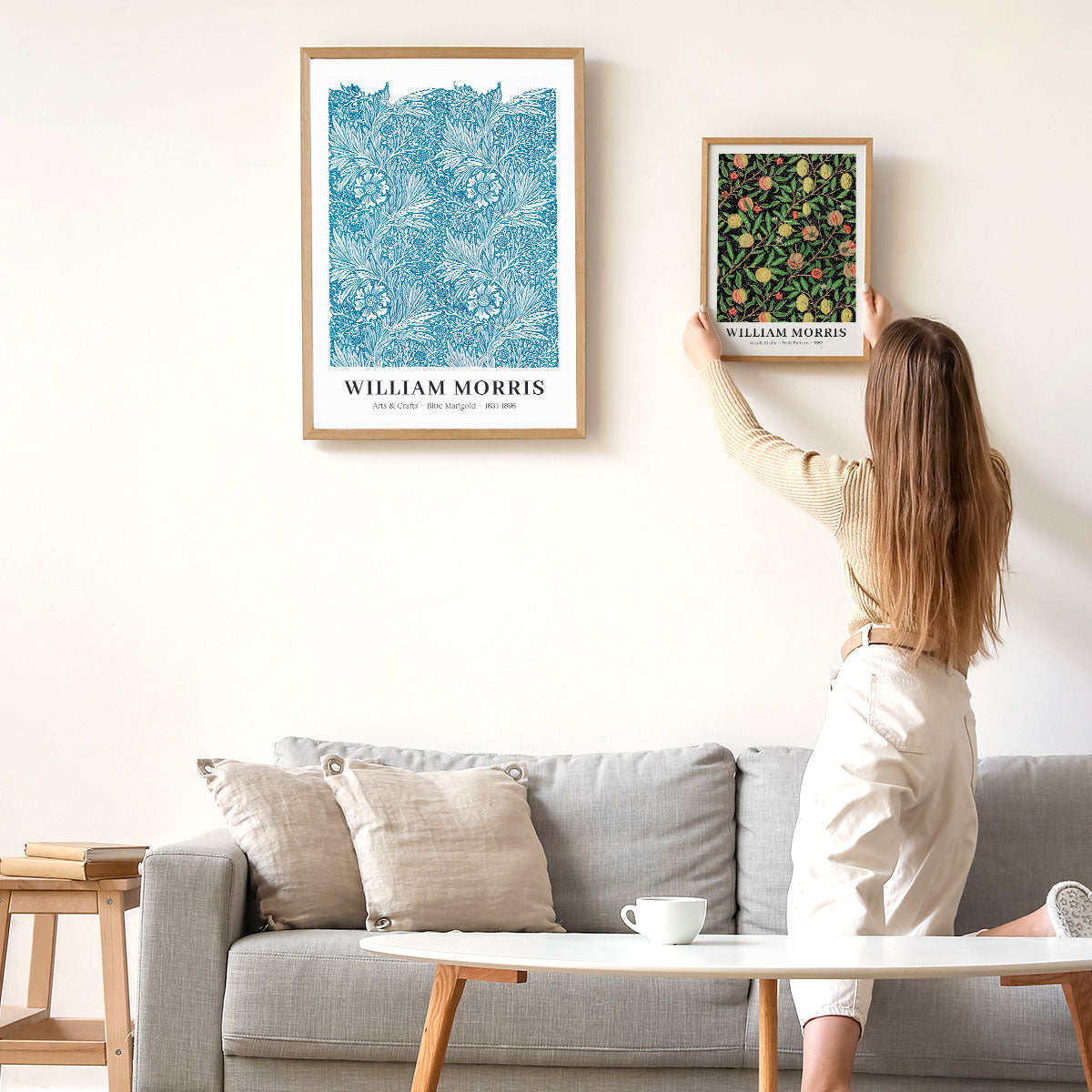 Kvinde hænger William Morris-plakater op på en væg over en grå sofa.