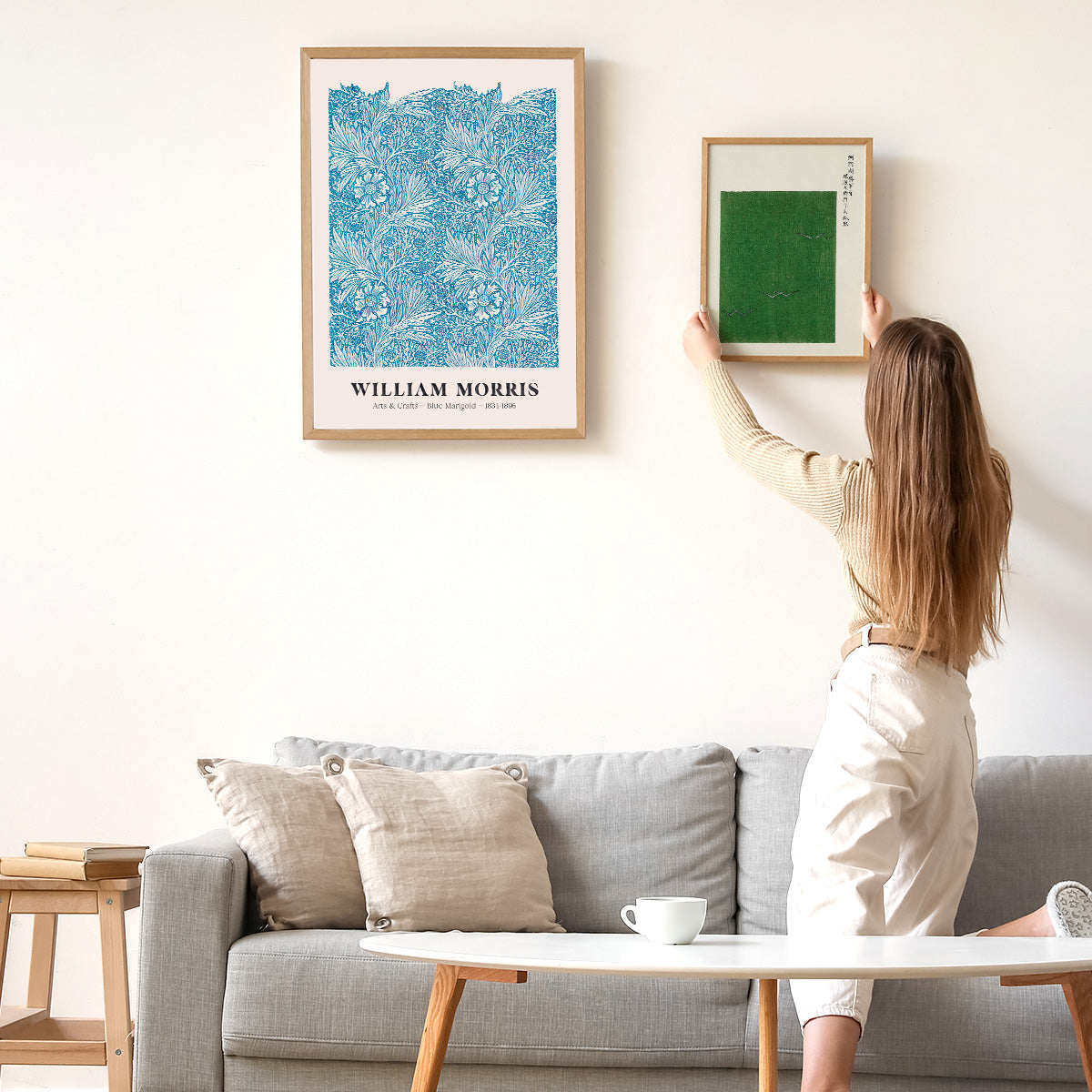Kvinde hænger to plakater op på en hvid væg over en grå sofa. Den ene plakat er en William Morris-design med blå blomstermønster.