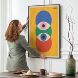 Kvinde hænger en Bauhaus-plakat op på en væg i en stue. Plakaten har et geometrisk design med cirkler i blå, grøn, rød og pink på en gul baggrund.