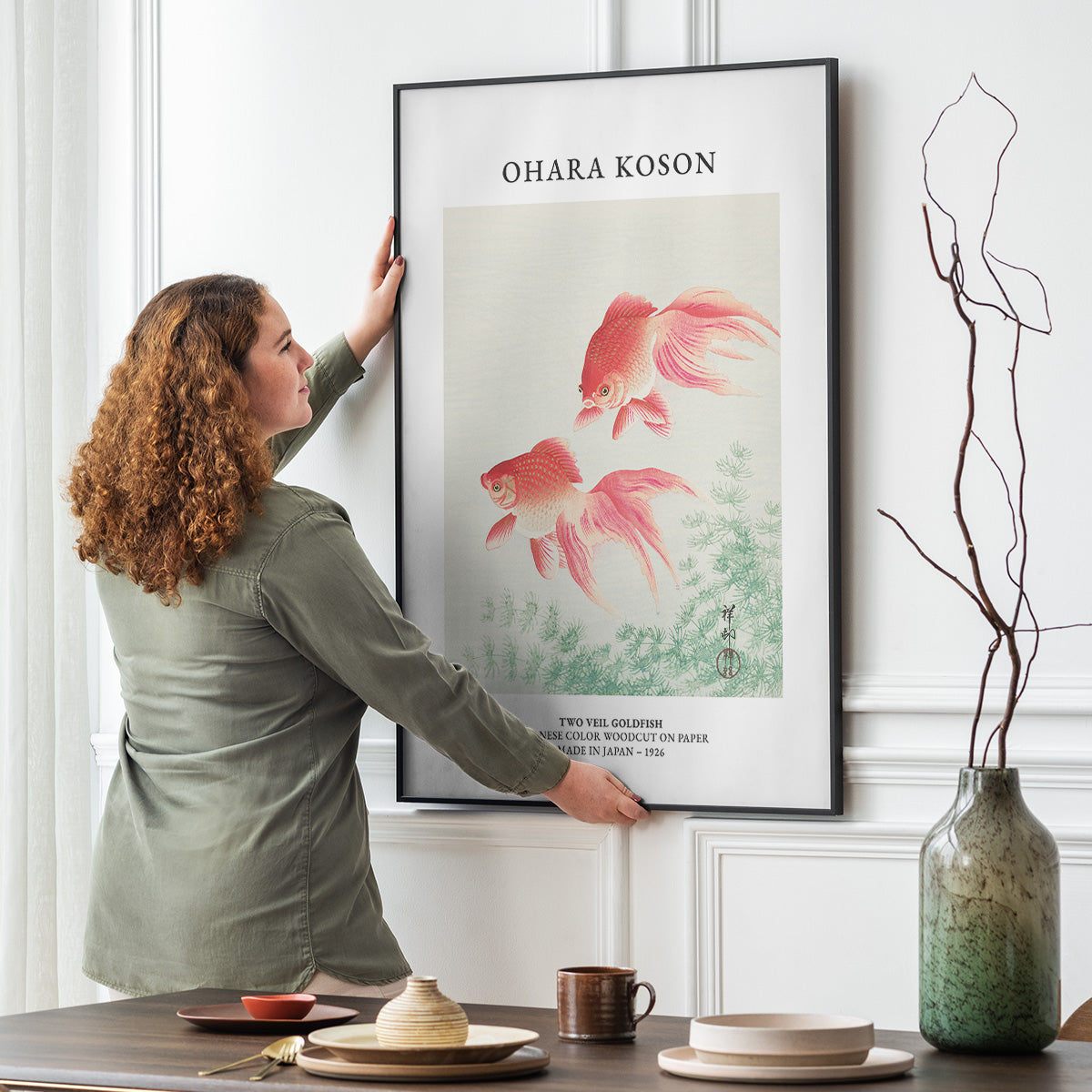 Kvinde hænger en plakat af Ohara Koson med guldfisk op på en væg i en stue.