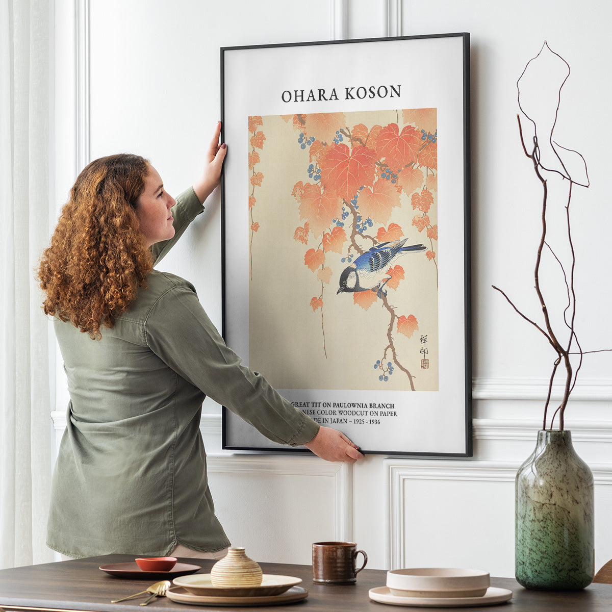 En person hænger en plakat op på en væg. Plakaten viser et kunstværk af Ohara Koson med en fugl og blomster.