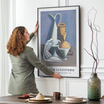 Kvinde hænger en plakat op på en væg i en stue. Plakaten viser et stilleben med vaser og frugter.