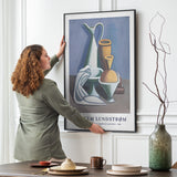 Kvinde hænger en plakat op på en væg i en stue. Plakaten viser et stilleben med vaser og frugter.