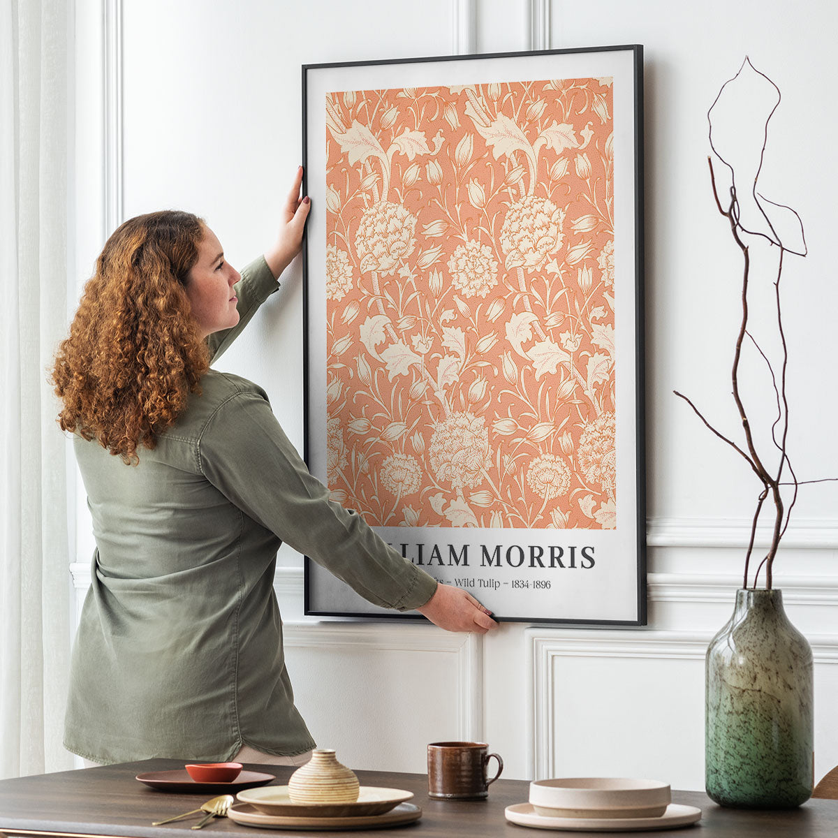 Kvinde hænger en William Morris plakat med titlen "Wild Tulip" op på en væg i en stilfuld stue.