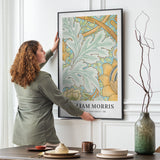 Kvinde hænger en William Morris plakat op på en væg i en stue. Plakaten viser et botanisk mønster i grønne og gule nuancer.