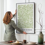 Kvinde hænger en plakat op på en væg. Plakaten viser et grønt bladmønster og har teksten "William Morris - Willow Leaves - 1873" nederst.