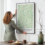 Kvinde hænger en plakat med et grønt, bladmønster på en væg. Plakaten er af William Morris og viser titlen "Willow Bough".