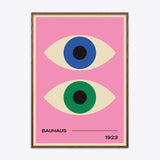 Plakat med to stiliserede øjne i blå og grøn på en lyserød baggrund, med teksten "Bauhaus 1923" nederst, indrammet i mørk naturtræsramme.