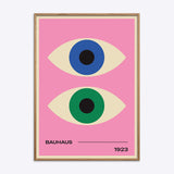 Bauhaus plakat med to stiliserede øjne i blå og grøn på en lyserød baggrund, indrammet i naturtræ.
