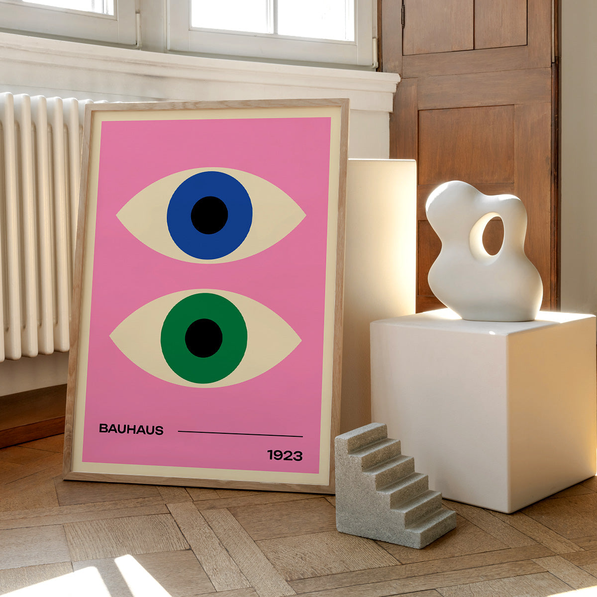 Bauhaus plakat med to øjne i forskellige farver på lyserød baggrund, placeret på et trægulv ved siden af moderne skulpturer.