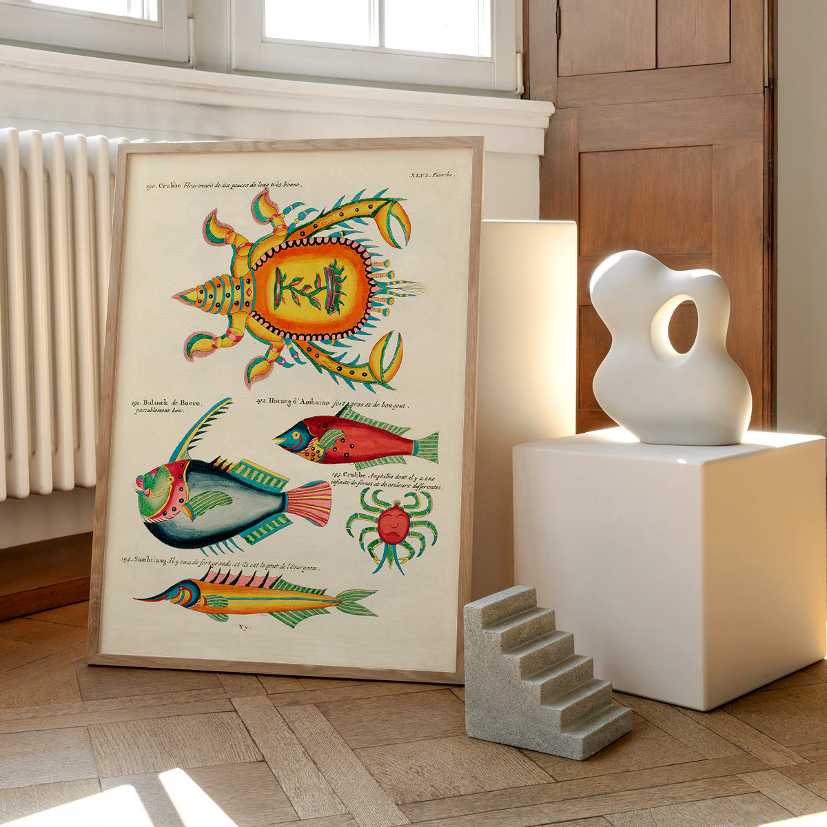 Plakat med farverige illustrationer af krabber og fisk, placeret på et gulv ved siden af moderne skulpturer og en radiator.