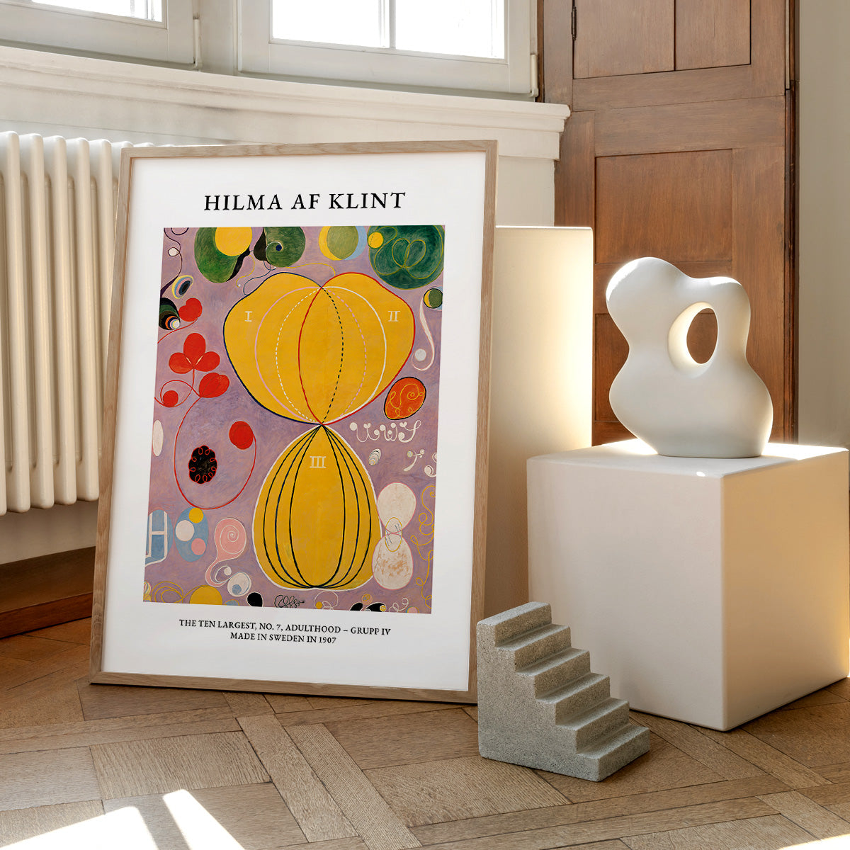 Plakat af Hilma af Klint med titlen "Adulthood" placeret på et trægulv ved siden af en skulptur og en lille trappe.