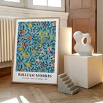 William Morris plakat med titlen "Fruit of Pomegranate" fra 1862, placeret på et trægulv ved siden af en moderne skulptur og en lille trappe.