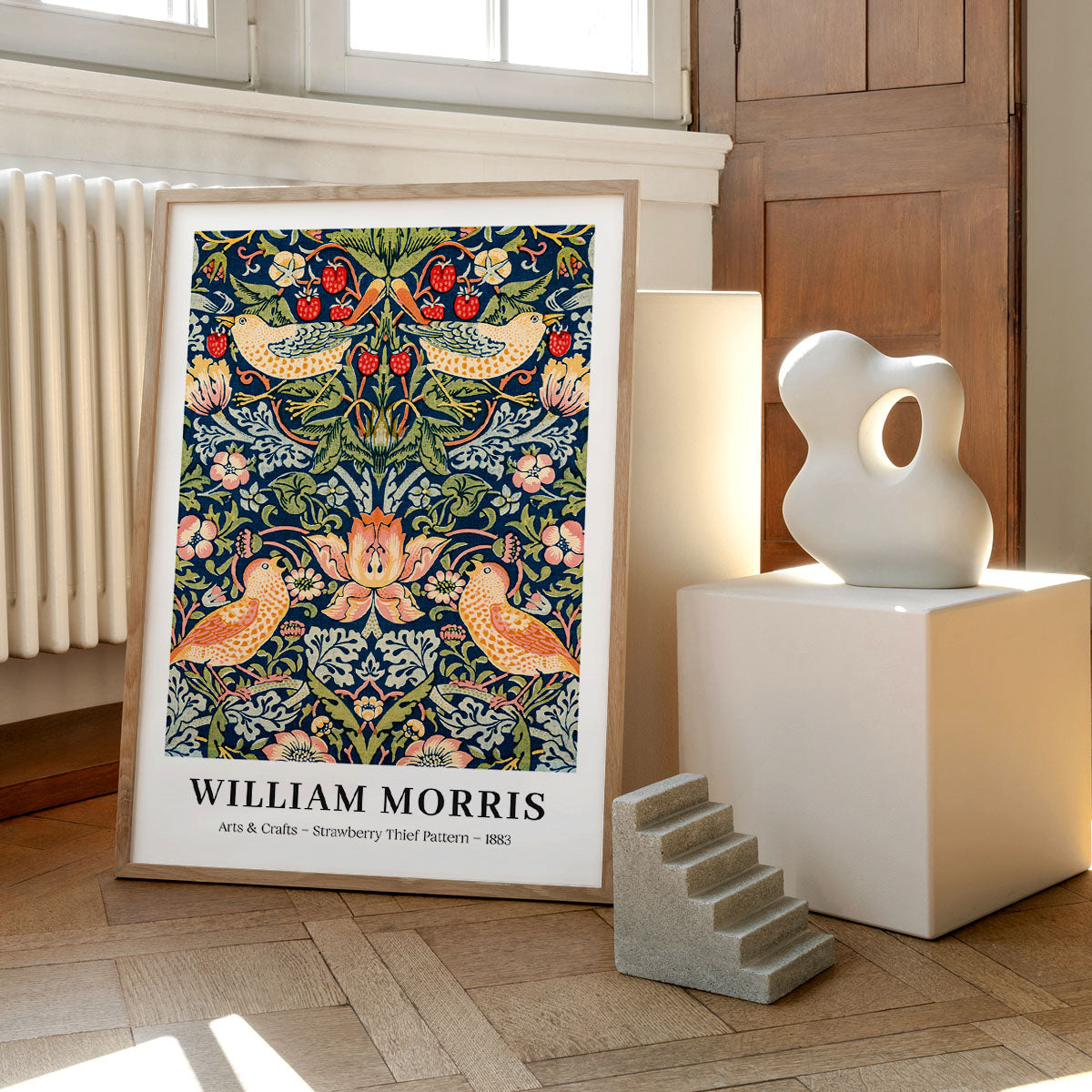 Plakat af William Morris med titlen "Strawberry Thief Pattern - 1883" placeret på et trægulv ved siden af en moderne skulptur og en lille trappe.