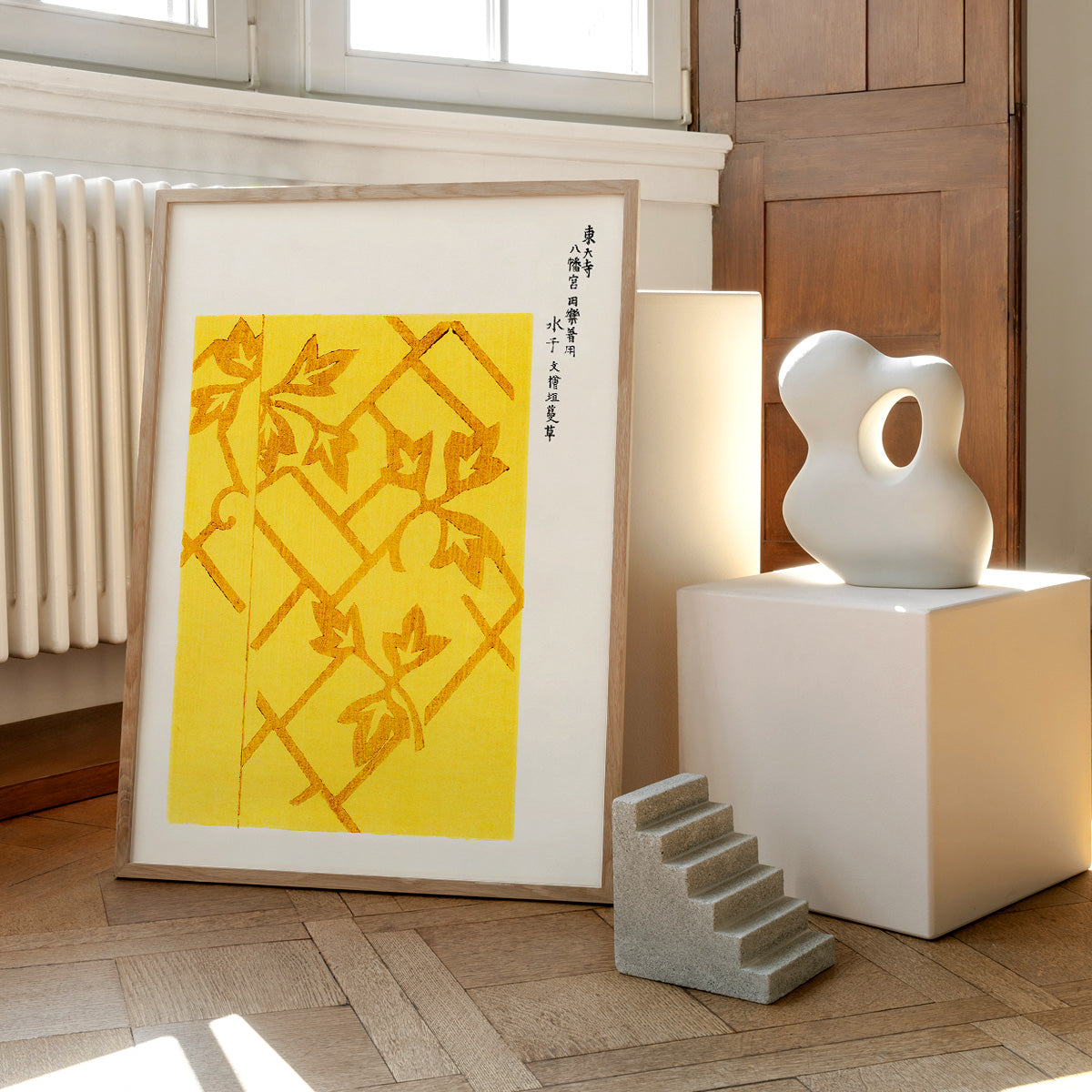 Plakat med gult og brunt bladmønster, placeret på et trægulv ved siden af en moderne skulptur og en lille trappeformet dekoration.