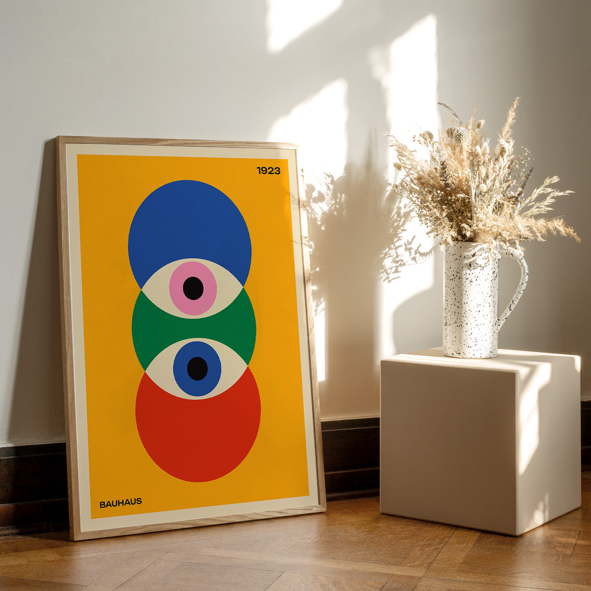 Bauhaus plakat med farverige geometriske former, placeret på et trægulv ved siden af en vase med tørrede blomster.