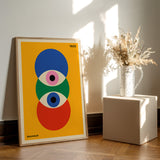 Bauhaus plakat med farverige geometriske former, placeret på et trægulv ved siden af en vase med tørrede blomster.