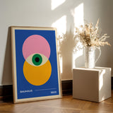 Plakat med Bauhaus-design fra 1923, viser geometriske former i blå, pink, gul og grøn, placeret på et trægulv ved siden af en vase med tørrede blomster.