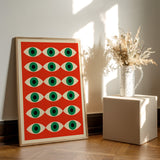 Plakat med et geometrisk mønster af øjne på en rød baggrund, placeret på et gulv ved siden af en vase med tørrede blomster.