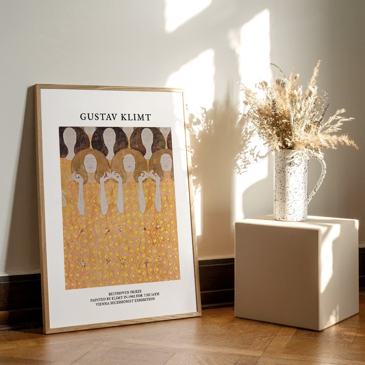 Gustav Klimt plakat med kunstværket "Beethoven Frieze" placeret på et trægulv ved siden af en vase med tørrede blomster.