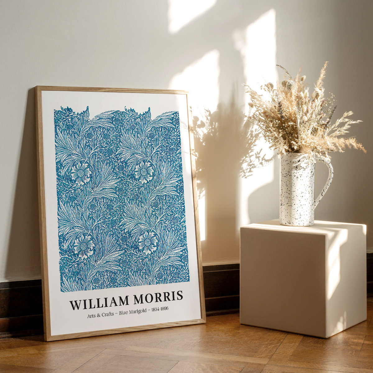 Plakat af William Morris med titlen "Blue Marigold" placeret på et trægulv ved siden af en vase med tørrede blomster.