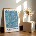 Plakat af William Morris med titlen "Blue Marigold" placeret på et trægulv ved siden af en vase med tørrede blomster.