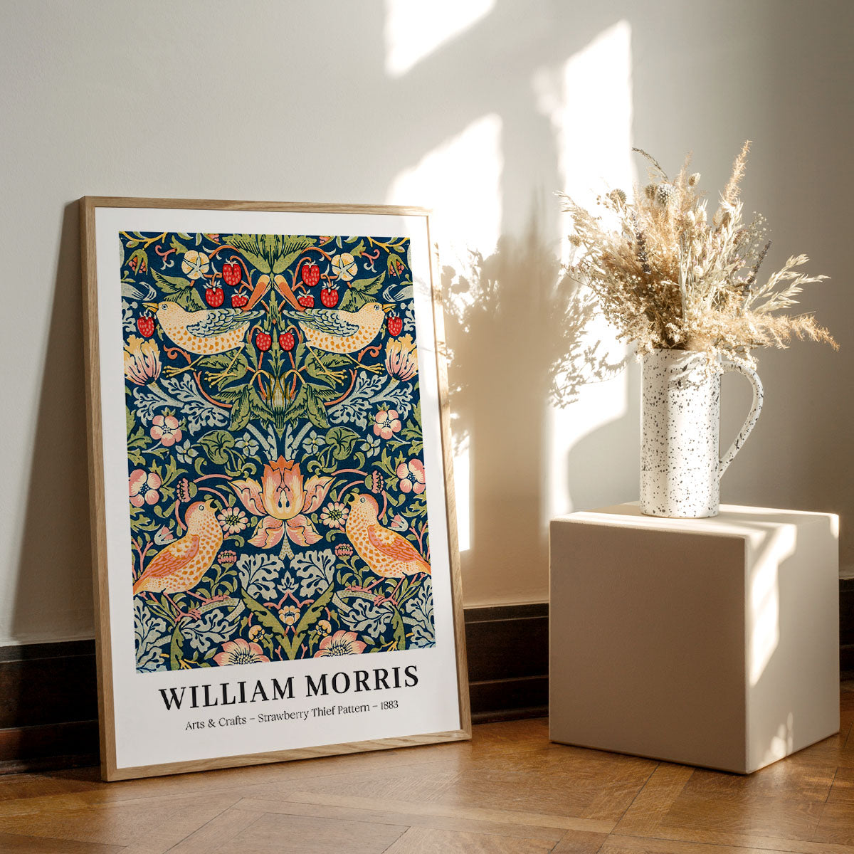 William Morris plakat med "Strawberry Thief Pattern" fra 1883, placeret på et trægulv ved siden af en vase med tørrede blomster.