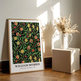 William Morris plakat med frugt- og bladmønster, placeret på et trægulv ved siden af en vase med tørrede blomster.