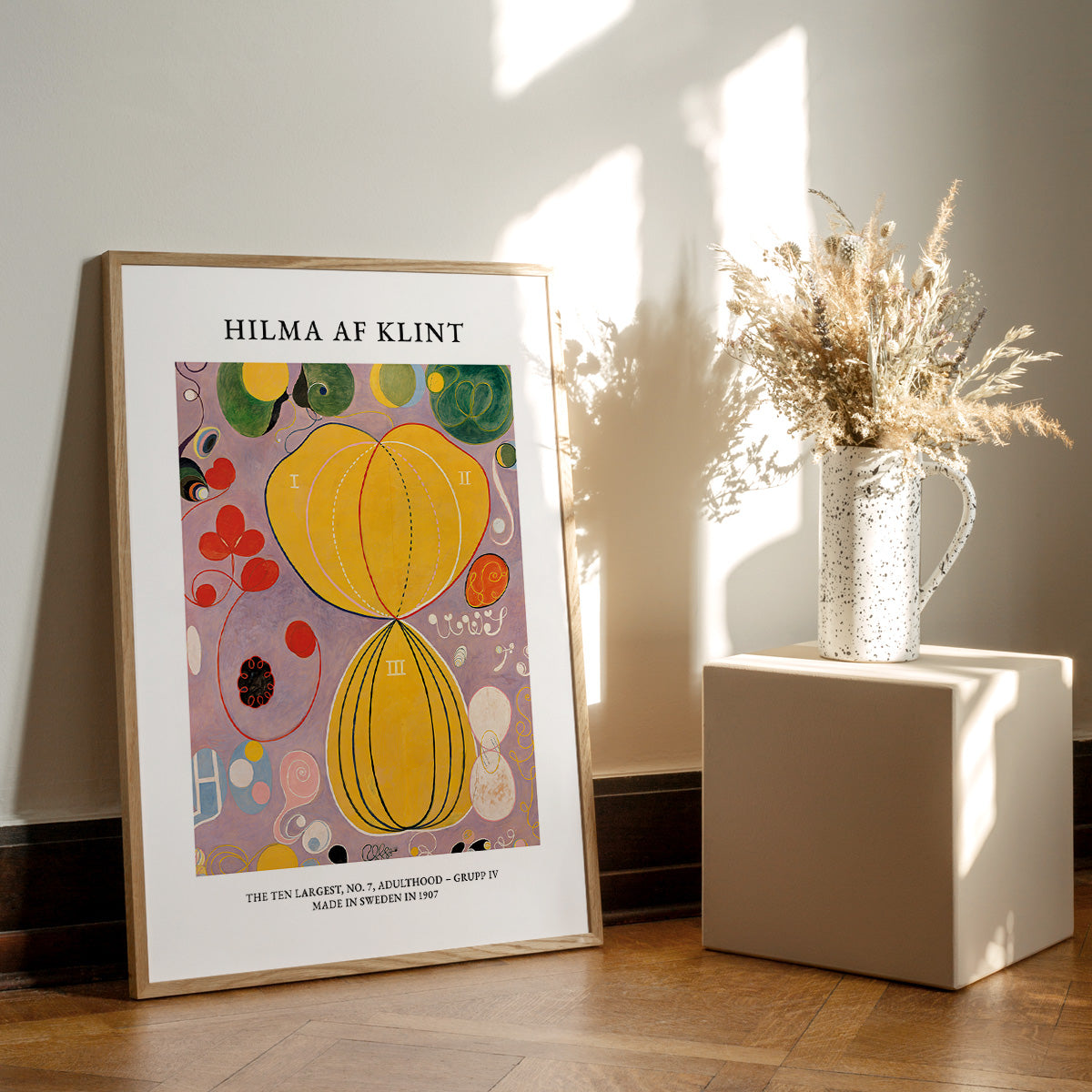 Plakat af Hilma af Klint med titlen "Adulthood" fra serien "The Ten Largest". Plakaten står på et trægulv ved siden af en vase med tørrede blomster.