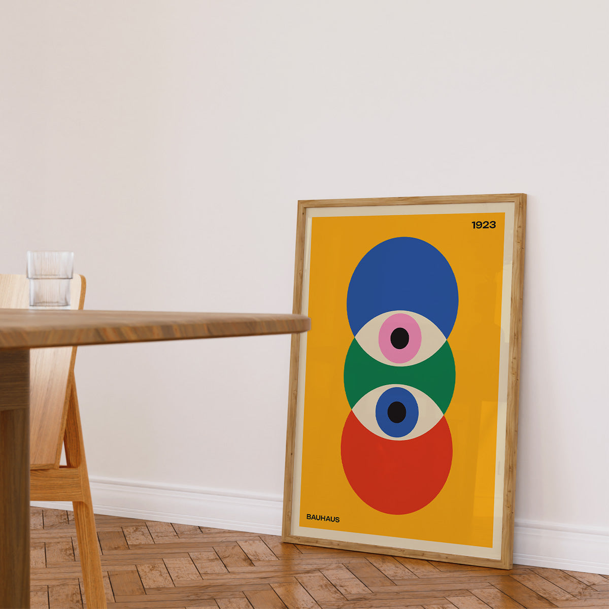 Bauhaus plakat med farverige cirkler, placeret i en træramme på gulvet ved siden af et træbord i en stue.