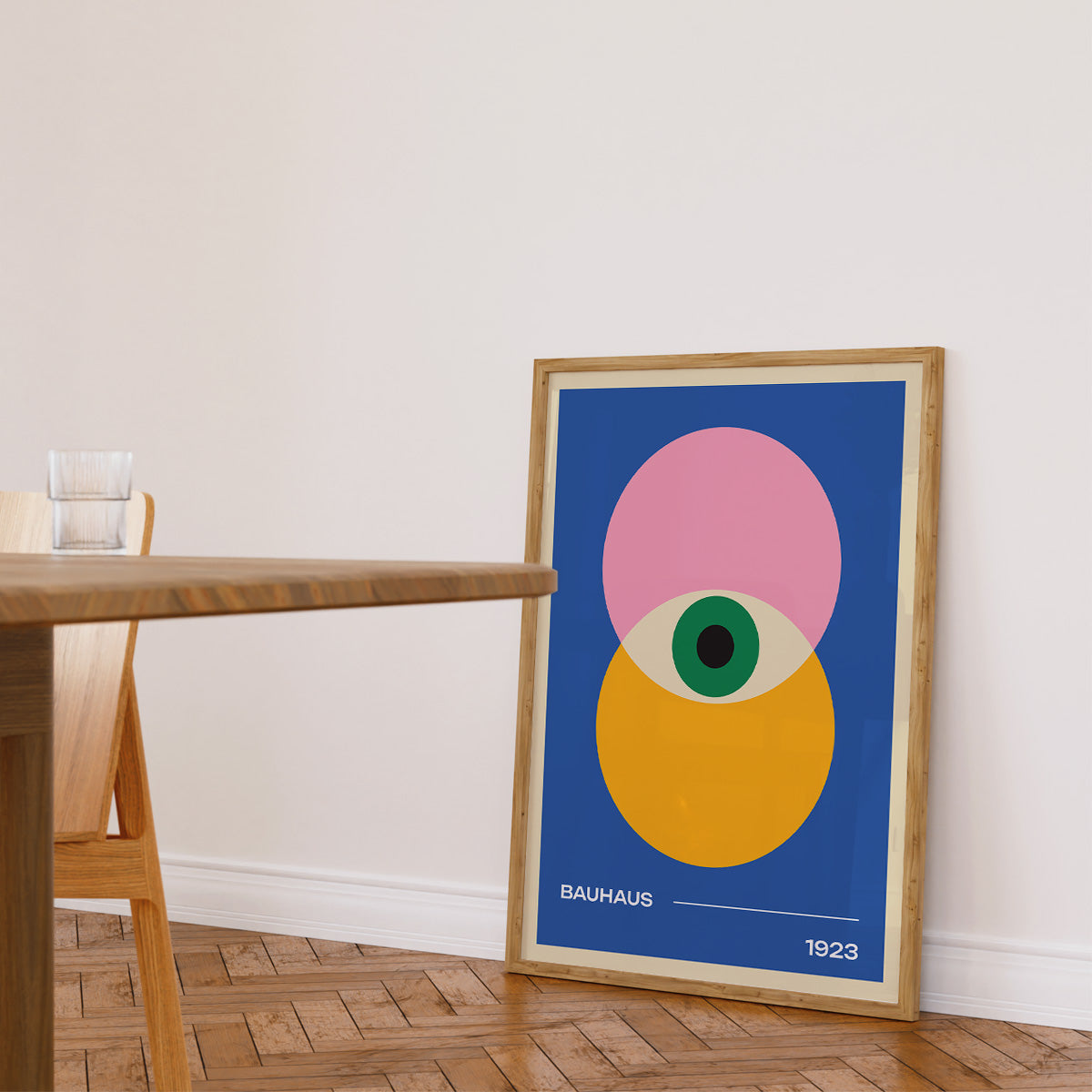 Plakat med Bauhaus-design fra 1923, viser geometriske former i pink, gul og grøn på en blå baggrund, indrammet og placeret på gulvet ved siden af et træbord i en lys stue.