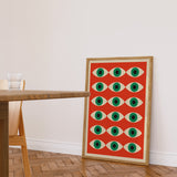 Plakat med geometrisk øjenmønster i røde og grønne farver, placeret på gulvet i en moderne stue.