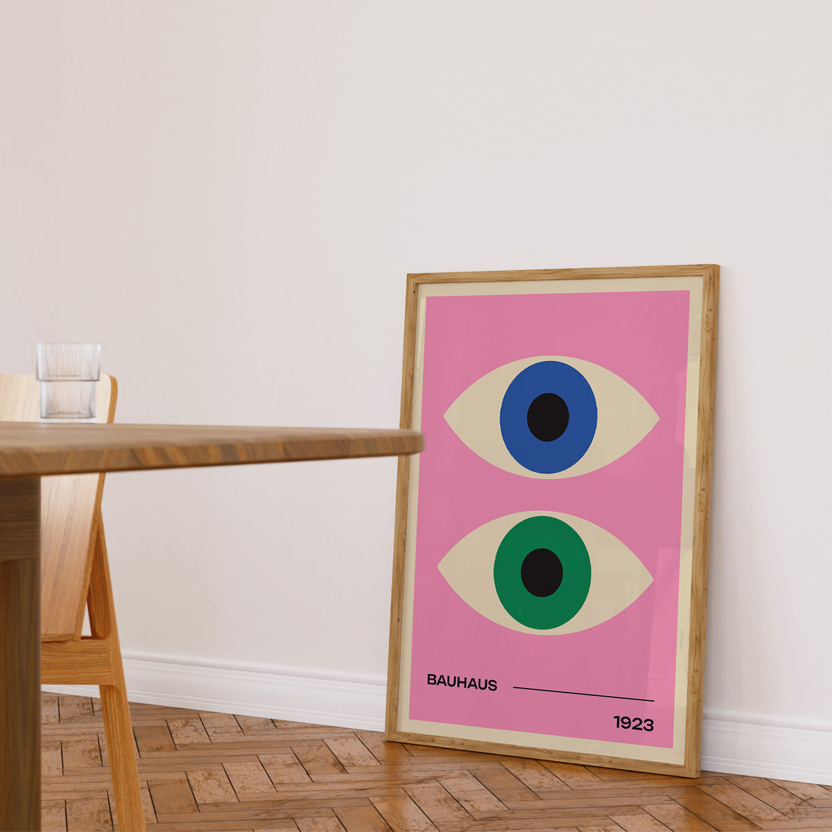 Bauhaus plakat med to øjne i forskellige farver på en lyserød baggrund, indrammet og placeret på gulvet ved siden af et træbord i en stue.