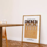 Gustav Klimt plakat i ramme, placeret på gulvet i en stue ved siden af et træbord og en stol.