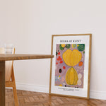 Plakat af Hilma af Klint med titlen "Adulthood" i en ramme, placeret på et trægulv ved siden af et bord.
