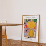 Plakat af Hilma af Klint med titlen "Adulthood" i en ramme, placeret på et trægulv ved siden af et bord.