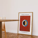 Plakat af Hilma af Klint med geometrisk design, placeret på gulvet i en stue ved siden af et træbord.