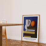 Plakat af Vilhelm Lundstrøm med stillebenmotiv, der viser en kande, en bog og en appelsin, placeret på gulvet i en moderne stue.