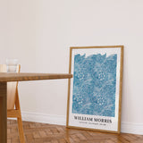 William Morris plakat med blå blomstermønster, placeret på gulvet ved siden af et træbord i en stue.